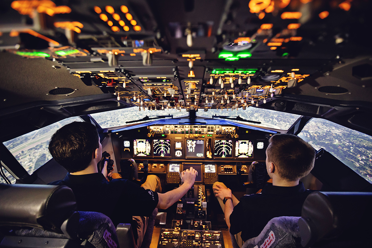 Two individuals sitting in the cockpit of an airplane simulator, operating controls and monitoring illuminated instrument panels, with a simulated aerial view visible through the windows.