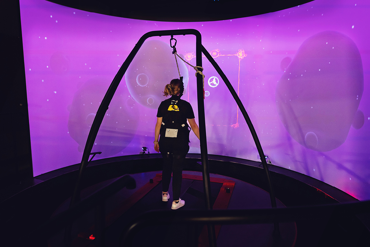 A person wearing a harness and motion sensors interacts with a biomechanics simulation in a lab setting, with a large curved screen displaying a purple virtual environment in the background.