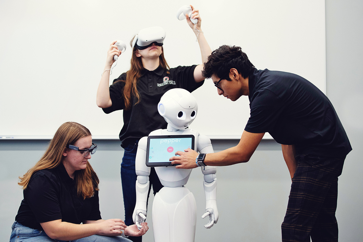 Three students interacting with a humanoid robot named Pepper; one student adjusts the robot's touchscreen, another crouches while examining its hand, and a third student wears a virtual reality headset, holding controllers in an immersive environment.