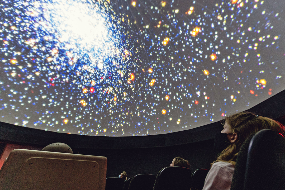 Visitors seated in a planetarium, gazing at a vibrant projection of a star-filled galaxy displayed across the domed ceiling.