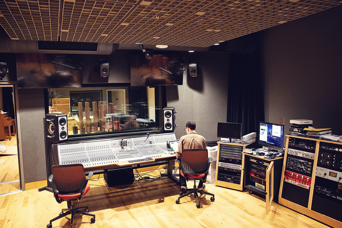 A technician seated at a large audio mixing console in a professional recording studio, with studio monitors, computer screens, and audio equipment surrounding the workspace, and a recording room visible through the glass window.