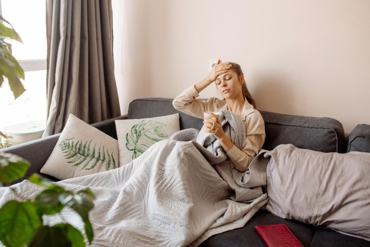 A woman relaxes on a couch with pillows and a blanket. She is hold a cup of tea in one hand and has her other hand on her forehead.