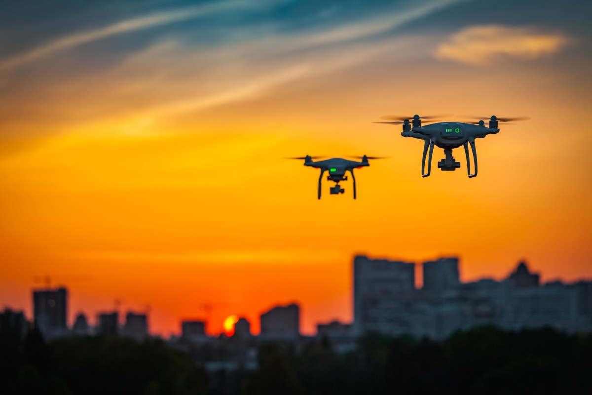 Two drones flying at sunset with a vibrant orange and blue sky in the background. The drones are equipped with cameras, hovering over a cityscape silhouette.