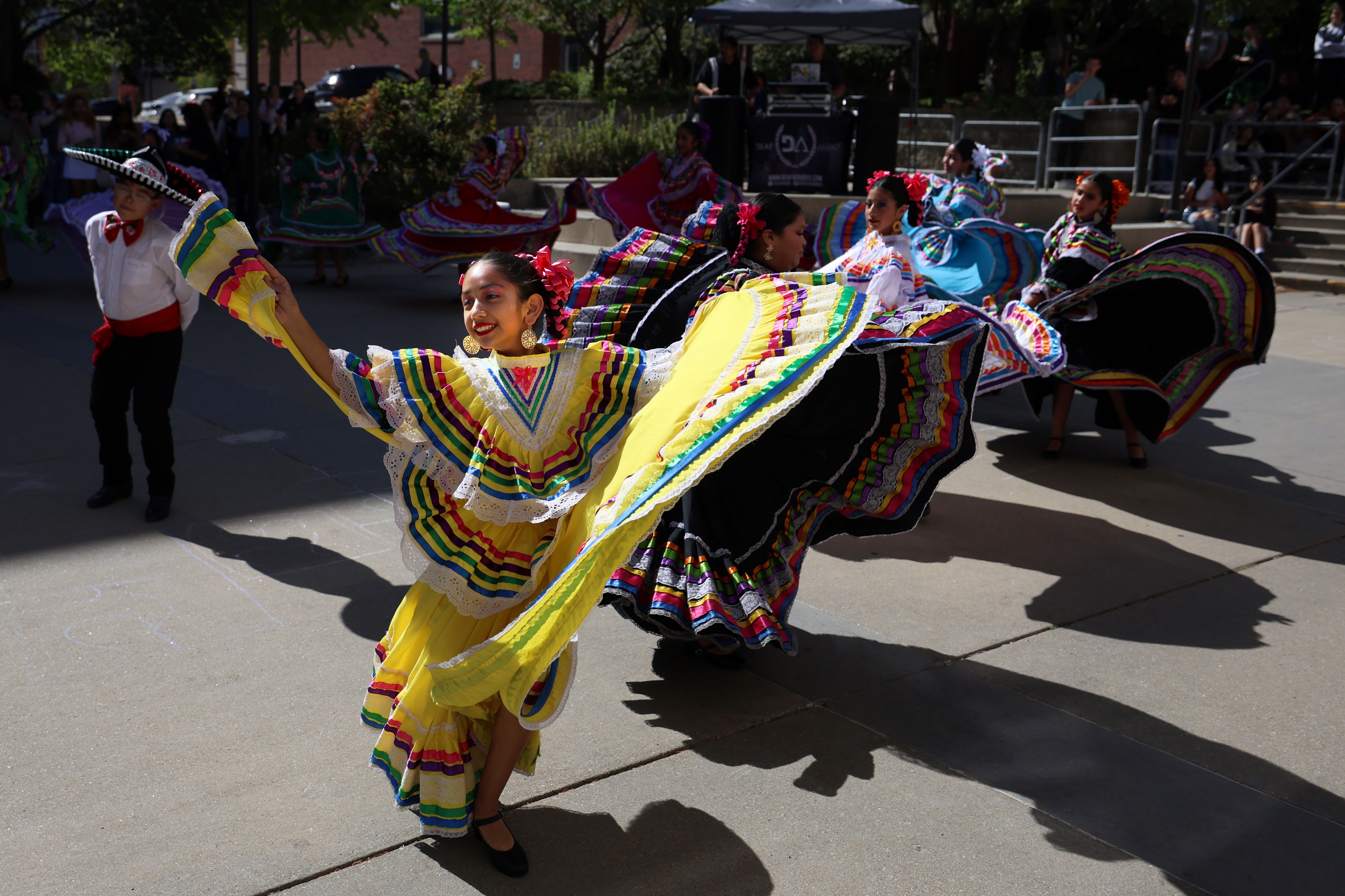 Dancers in colorful traditional Mexican attire perform at an outdoor event during Fiesta on the Plaza. A woman in a vibrant yellow dress is in the foreground, with others twirling in the background.