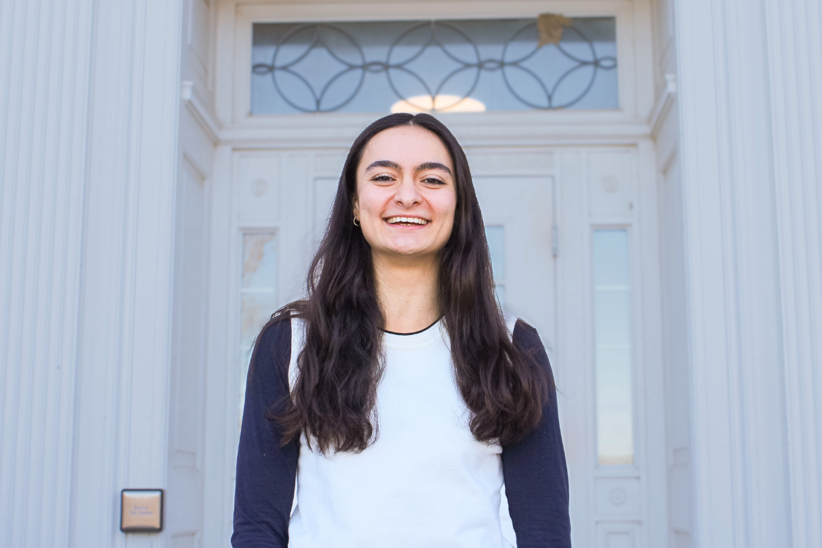 A woman with long dark hair smiles in front of a white building with decorative glass windows and ornate details.