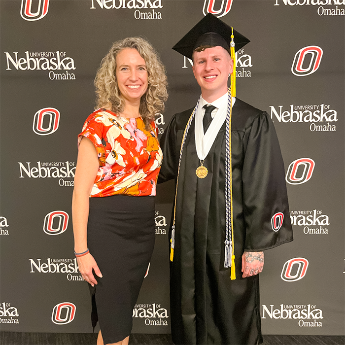 Aaron stands at commencement in a cap and gown, next to Dean Melanie Bloom. 