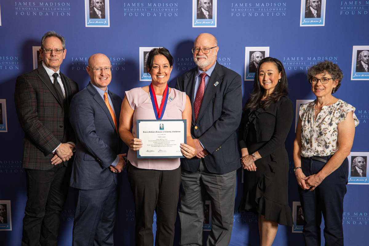 Six individuals posing together in front of a blue James Madison Memorial Fellowship Foundation backdrop. The central figure, a woman wearing a light purple top and a red medal around her neck, holds a certificate with a smile. The group includes three men and three women all dressed in professional attire.