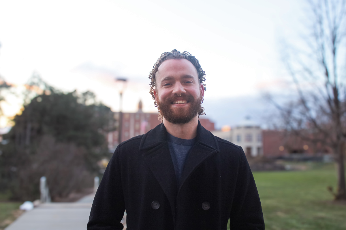 A man with curly hair and a beard smiles outdoors. He wears a black coat, and the background shows trees, buildings, and a cloudy sky.