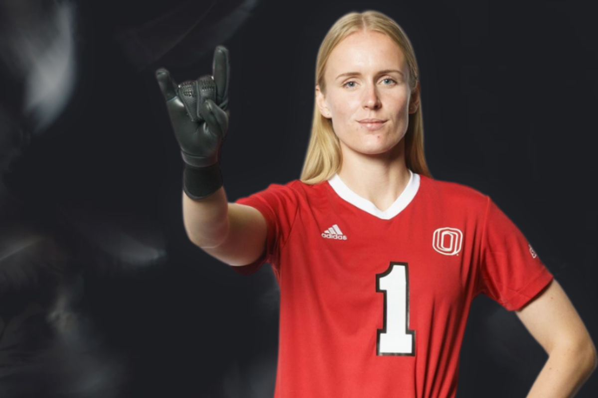 A woman with blonde hair wearing a red soccer jersey with the number 1 and the University of Omaha logo raises her gloved hand, making a confident gesture against a dark background.