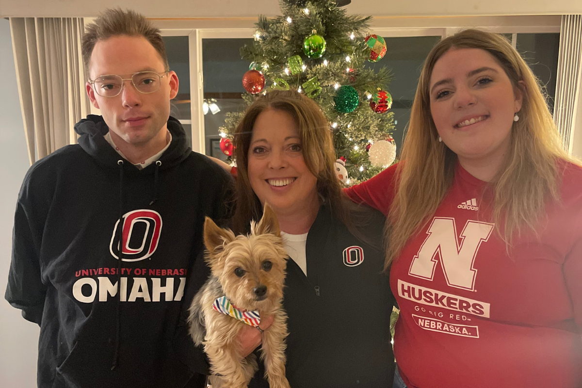 Three people pose indoors in front of a lit Christmas tree. The person in the middle holds a small dog with a colorful bow. They are dressed in University of Nebraska Omaha and Nebraska Huskers gear, smiling warmly.