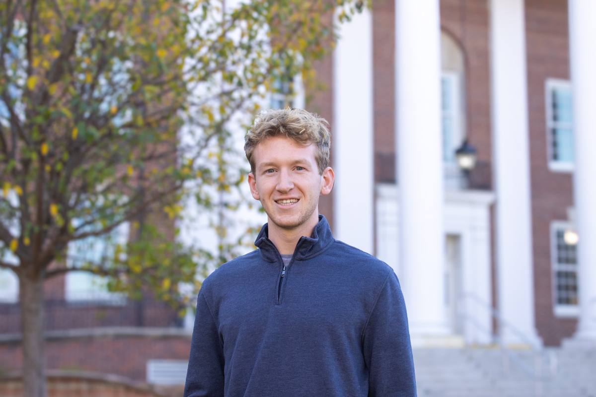 Simon Medinger smiles at the camera while standing outdoors in front of a brick building with white columns. He is wearing a navy-blue quarter-zip sweater, and the background features a tree with autumn leaves.