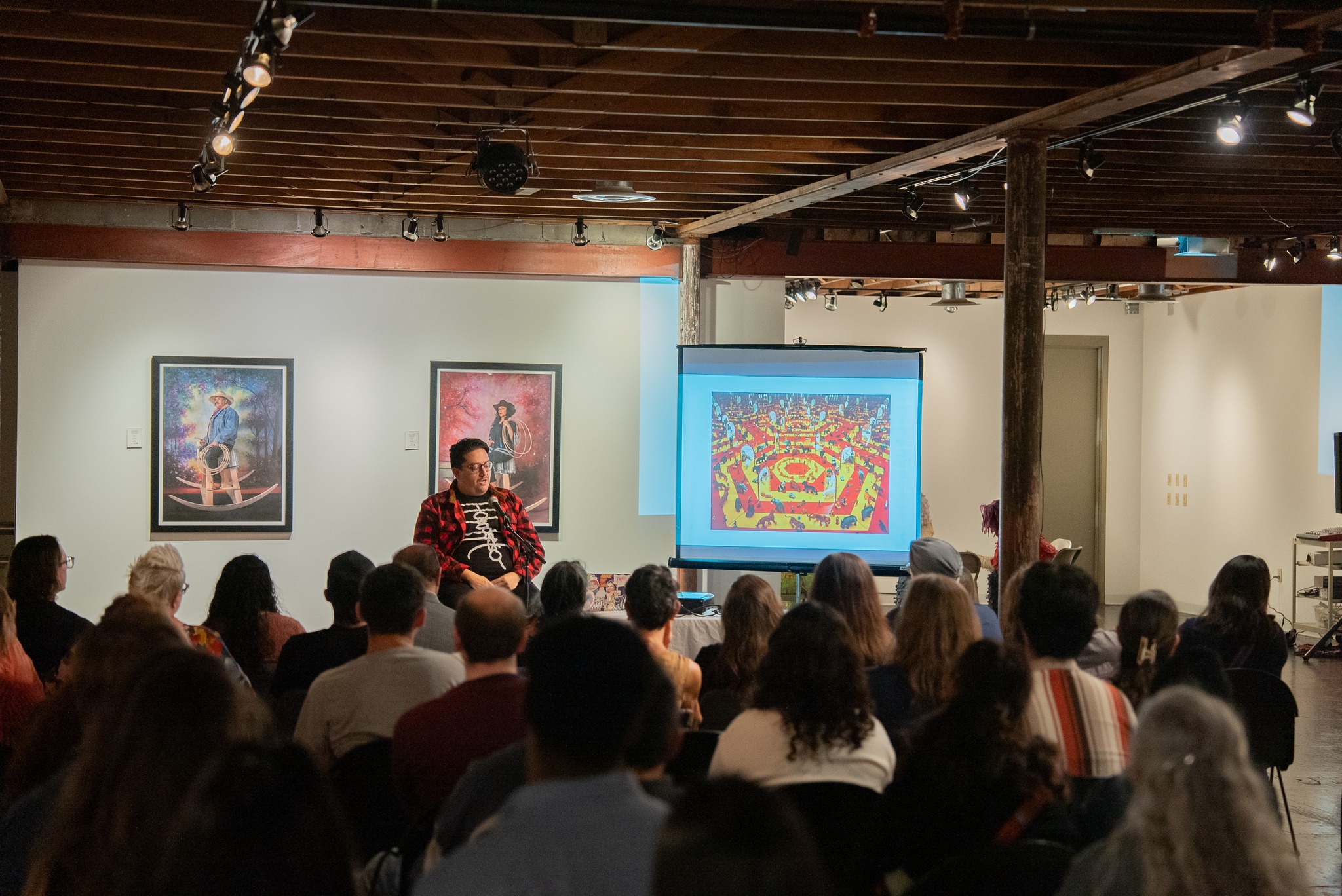 Justin Favela's presentation at the gallery infront of people sitting in chairs while he faces the people with a presentation beside him.