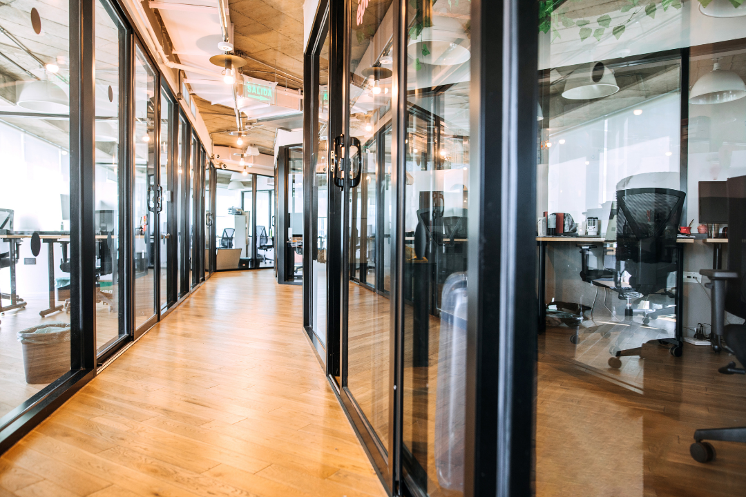 Bright modern office corridor with glass-walled rooms on both sides, wood floors, and desks and chairs visible inside.