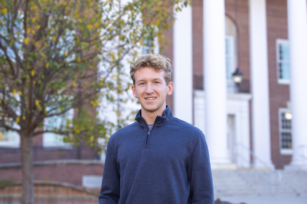 Simon smiles in front of a building with tall pillars and autumn leaves. He is wearing a navy sweater. 