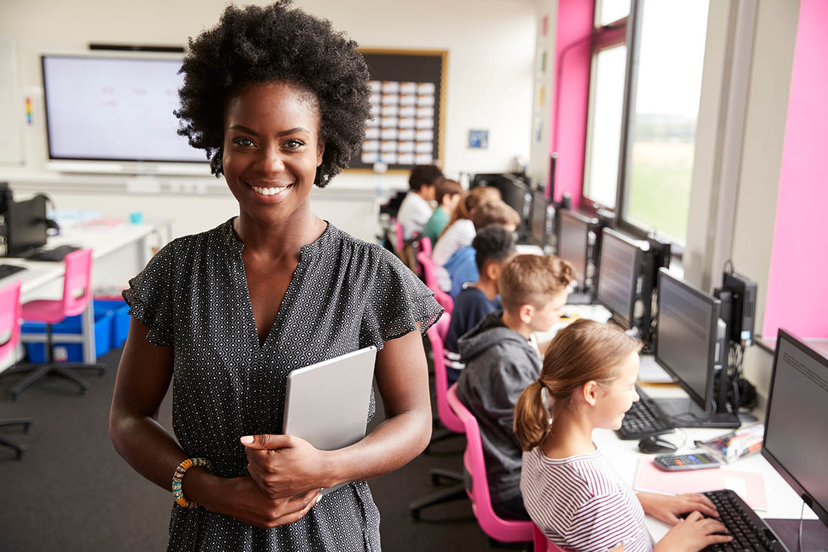 A teacher poses for a photo in her classroom; her students work on computers in the background