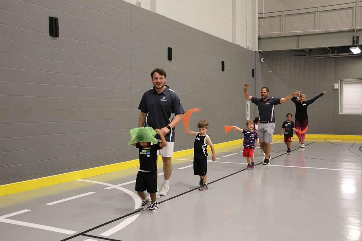 A group of kids walk on a line in a gym waving colorful flags; adults walk with them