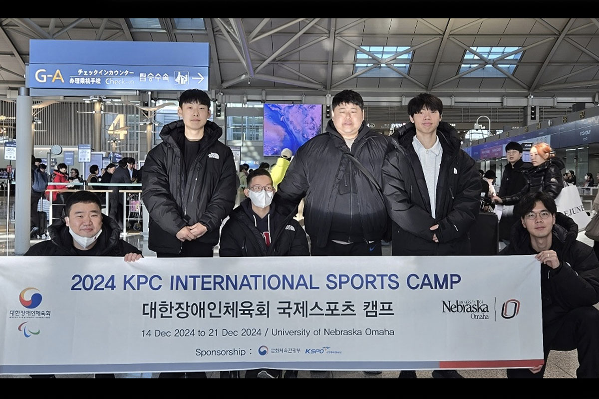 A group of people pose for a photo in an airport with a banner that reads International Sports Camp