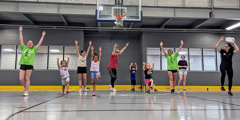 A group of adults and children stand in a line in a gym with their arms up