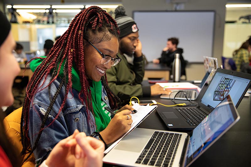 A woman smiling while working at a computer with other students in the background.