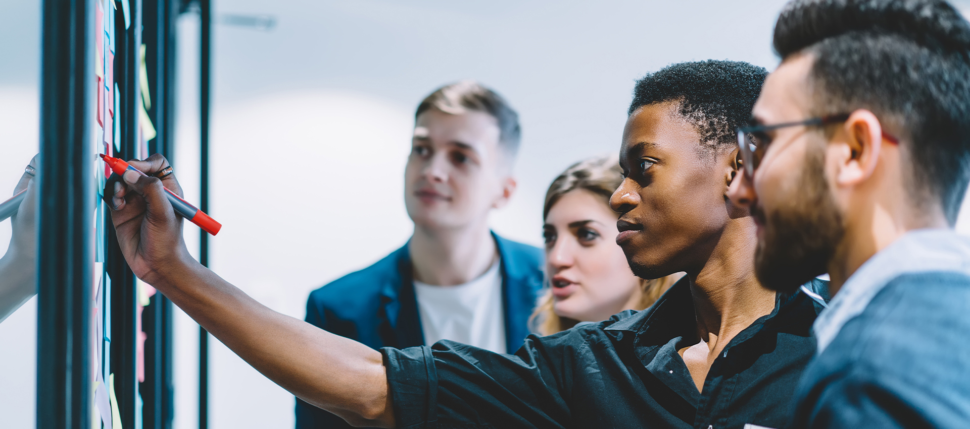 The image shows four management information systems students, a women and three men, standing in front of a white board scattered with multi-colored sticky notes. The African American man has his arm extended while holding a red dry erase marker while the others watch.