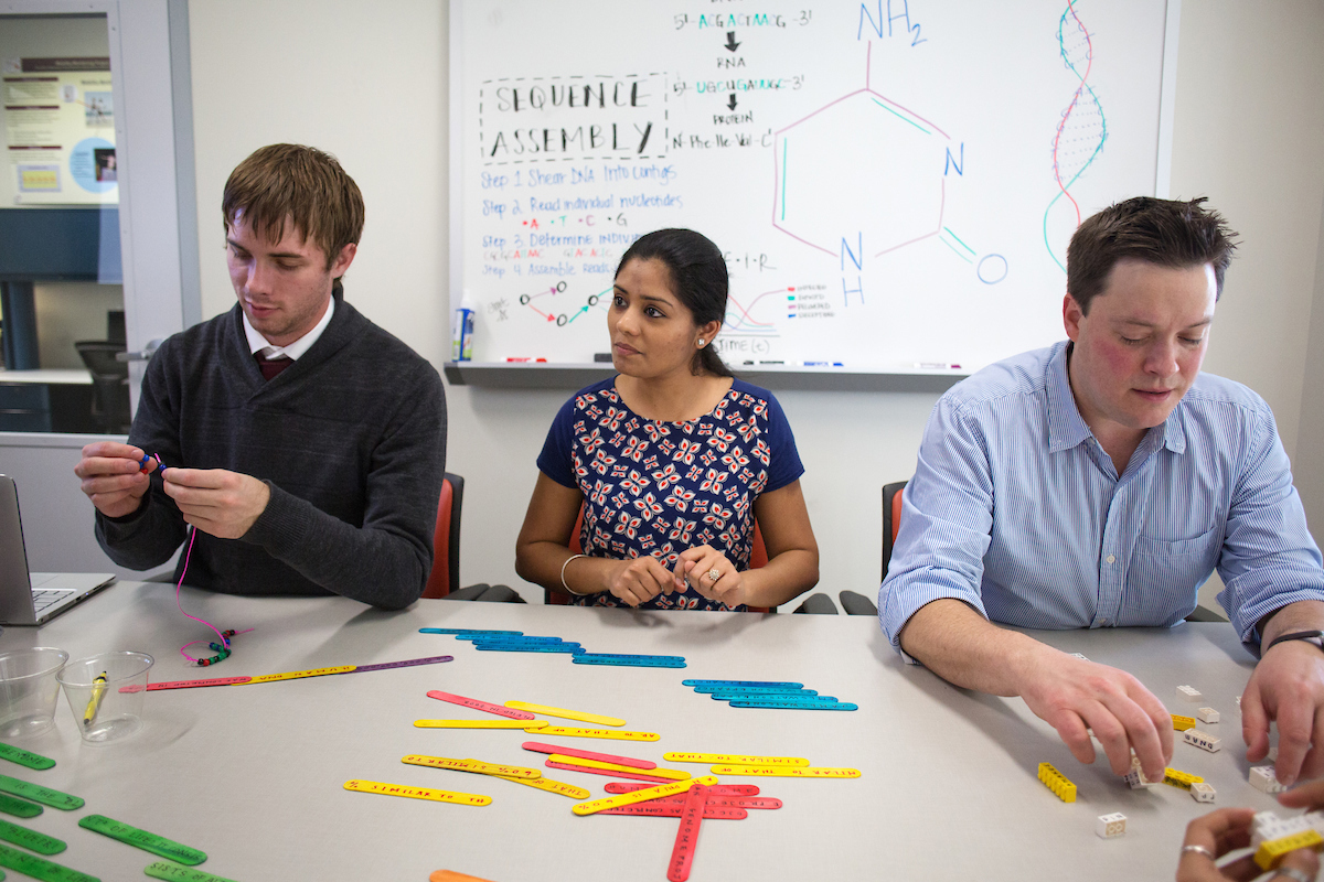 Three biomedical informatics student researchers working together in a laboratory, examining data on a computer screen. A microscope and other lab equipment are visible in the background. One student researcher is seated, using the computer, while the other stands beside, pointing at the screen.