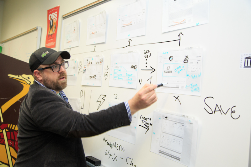 A man wearing glasses and a cap is standing next to a whiteboard filled with diagrams and notes. He is pointing at one part of the diagram while explaining something, with various paper prototypes pinned on the board.