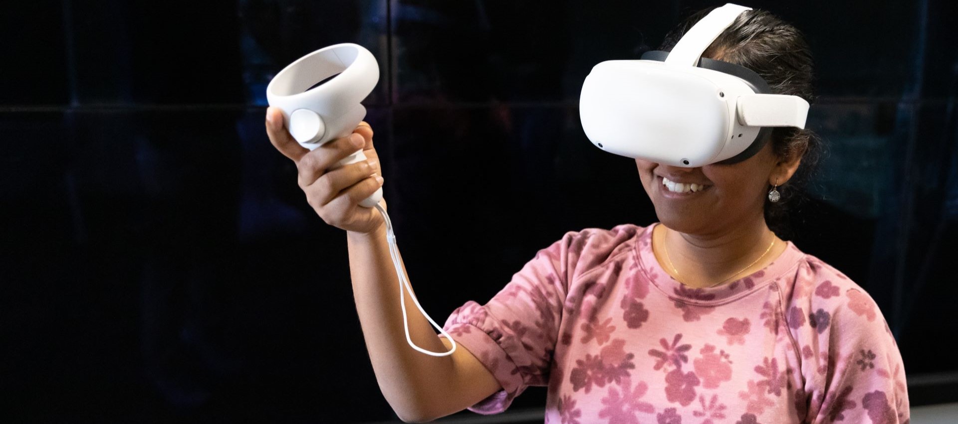 A woman is wearing a white virtual reality headset and holding a VR controller in her right hand. She is smiling and appears to be enjoying the VR experience. She is dressed in a pink floral-patterned top and is standing against a dark background.