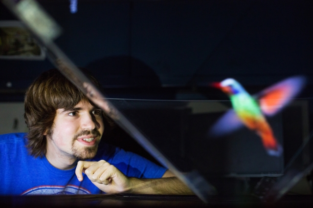 A young man is observing a hologram of a colorful hummingbird that appears to be floating in mid-air. He is smiling and seems fascinated by the holographic image.