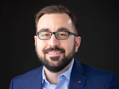 The image shows the Director of the School of Interdisciplinary Informatics, Matt Hale, a man with a friendly expression. He has short, neatly styled brown hair and a light beard. He is wearing glasses with dark, rectangular frames and a blue suit over a blue shirt. The background of the image is black, which highlights his features and professional attire. This portrait style is typical for corporate or professional profiles.