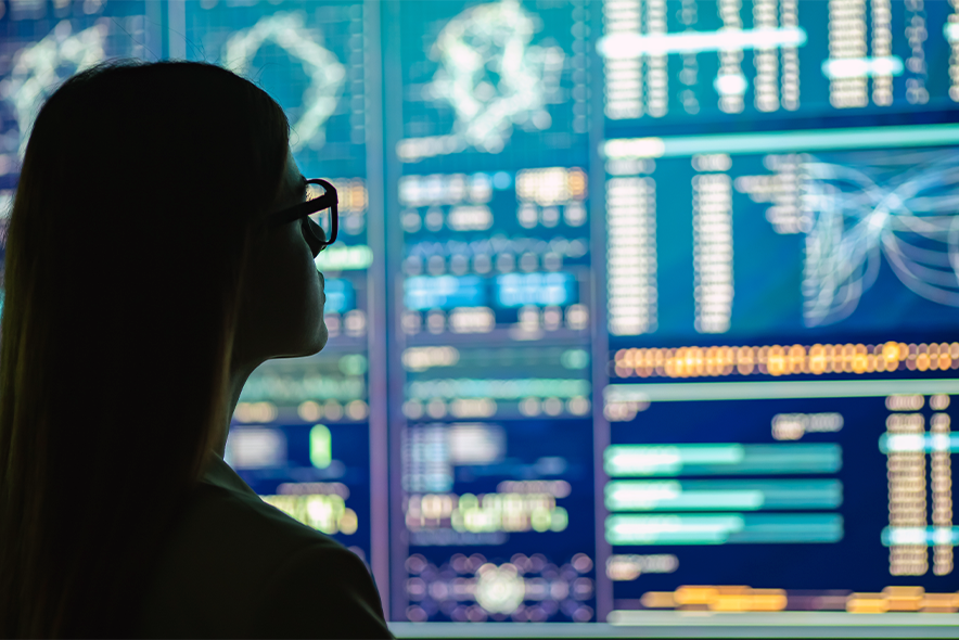 A businesswoman in glasses standing near a large digital display depicting several applications relating to data science.