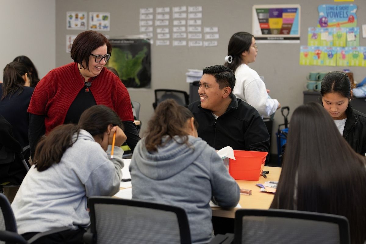 A teacher smiles and talks with a student in a classroom while several other students work at their desks.