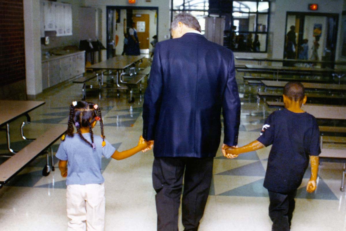 a man with two students, one holding each of his hands, walking down a hallway with their backs facing the camera