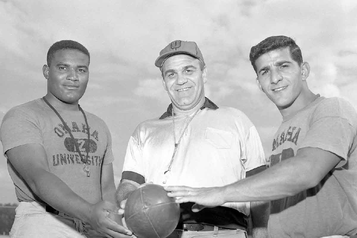 three Omaha University football coaches with their hands on a football looking into the camera