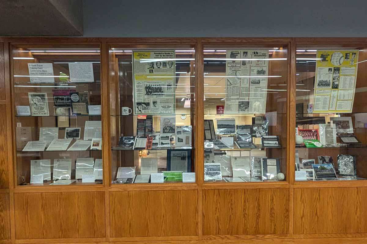 built in glass and wood display shelves. on the shelves there are photos , newspaper clippings, and other ephemera about women's softball at UNO