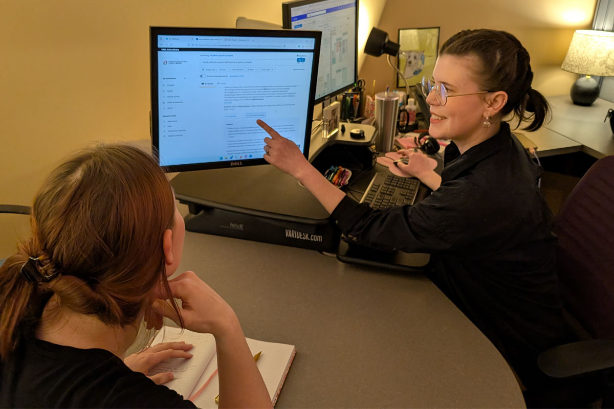 Two people in an office discussing content on a computer screen. One person is wearing glasses and a black top while pointing at the computer screen. The other person is facing the computer screen and appears to be listening.
