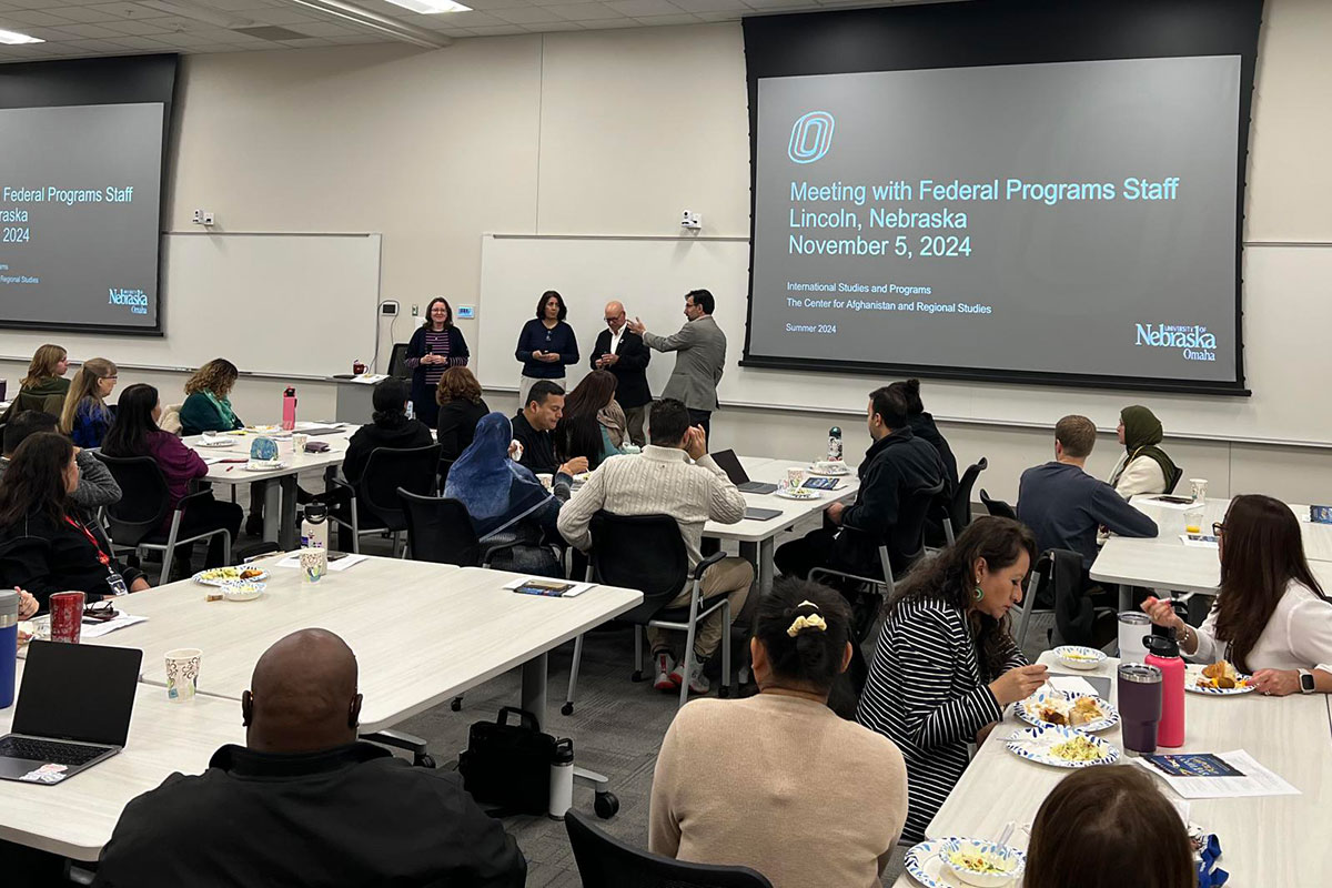 Large classroom filled with working professionals sitting around tables. Digital displays featuring a powerpoint presentation appear in the back ground, and 4 people presenting are at the front of the room.