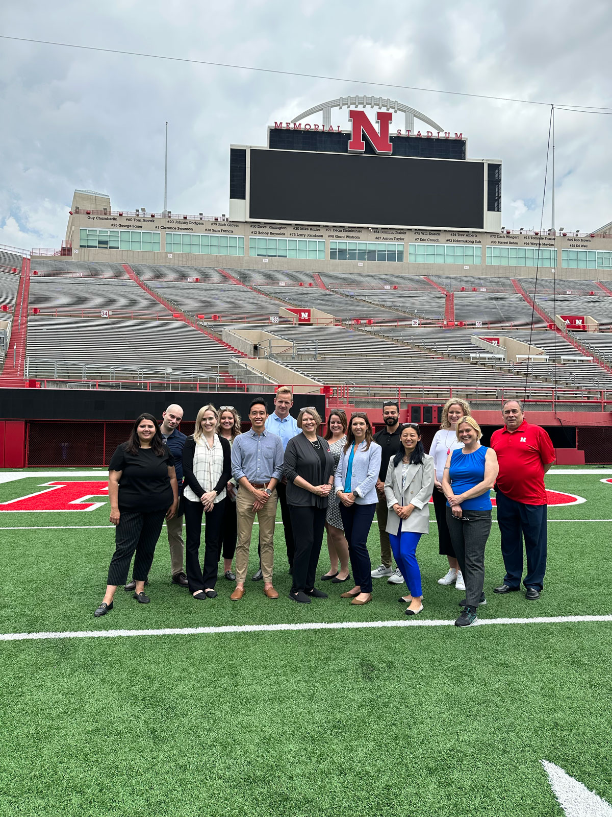 Group of people standing on a football field at a stadium with the Nebraska Cornhuskers logo in the background.