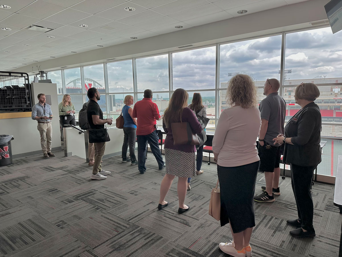A group of people standing in a modern conference room with large windows, looking out at a stadium view.