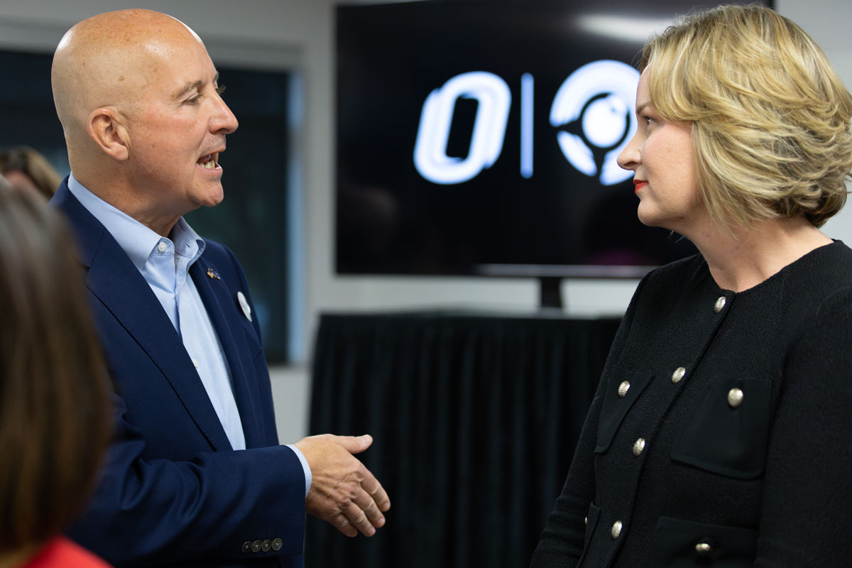 A closeup shot of Pete Ricketts, left, speaking with Gina Ligon, right. In the background between them is a black banner bearing the UNO O and the NCITE birdie.