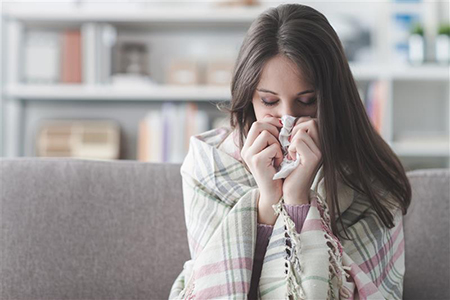 Sick young woman at home on the sofa with a cold, she is covering with a blanket and blowing her nose