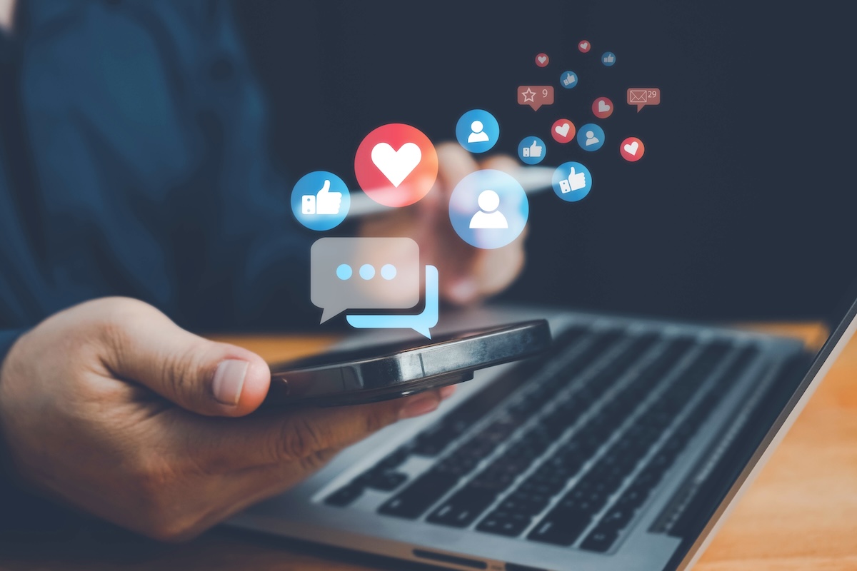 A person using a smartphone while working on a laptop, with digital social media icons, such as likes, hearts, comments, and follower symbols, floating above the phone, representing online engagement and interaction.