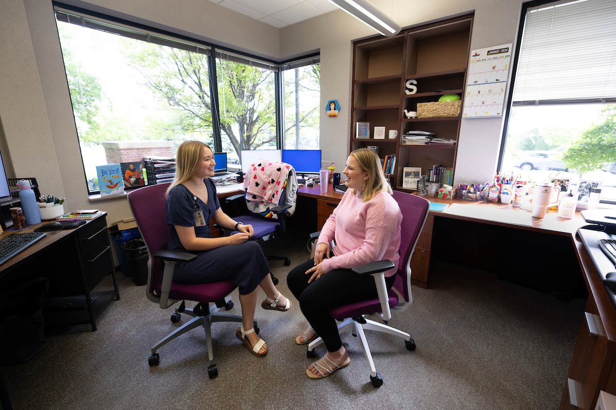 Claire Ramacciotti and Tess Willis sit across from each other in an office at Project Harmony, engaged in conversation, with a colorful and organized workspace in the background.