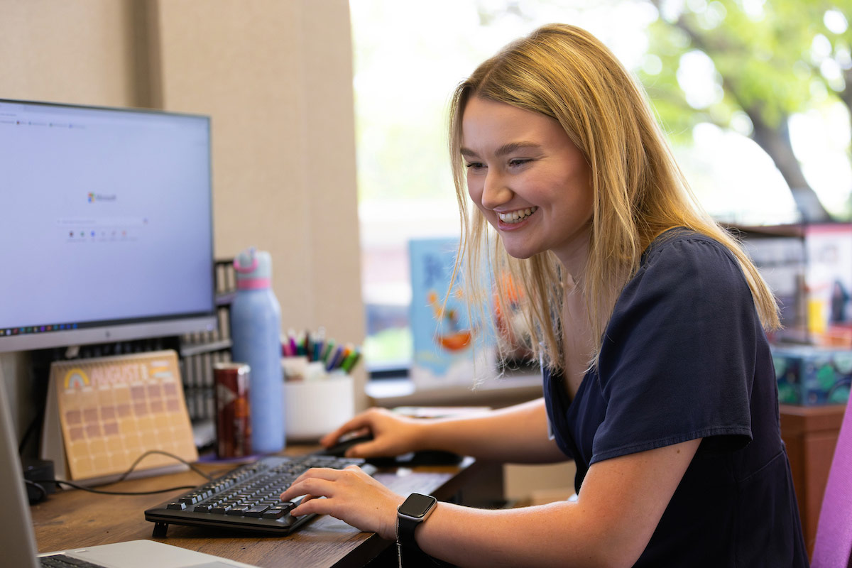 Claire Ramacciotti sits at her desk, smiling while typing on her keyboard, surrounded by colorful office supplies and a laptop, during her internship at Project Harmony.