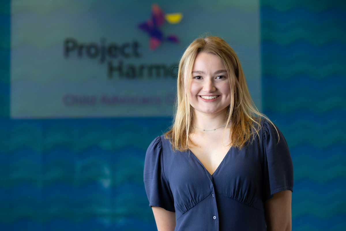 A smiling Claire Ramacciotti stands in front of the Project Harmony sign, dressed in a navy blue shirt, showcasing her internship in child advocacy.