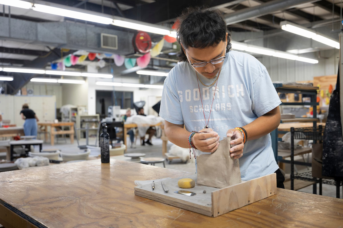 A student in a pottery class at the University of Nebraska at Omaha carefully molds a piece of clay on a work surface in a well-lit ceramics studio.