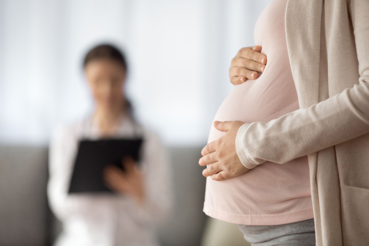 A close-up of a pregnant woman gently holding her belly with one hand, wearing a light pink top and beige cardigan. In the blurred background, a healthcare professional is holding a clipboard, appearing out of focus. The setting appears to be a medical or clinic environment.