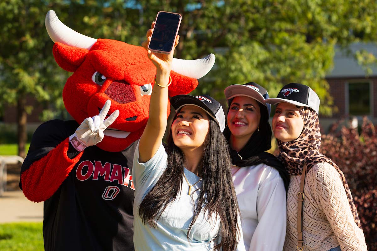 Durango, the UNO mascot, poses for a selfie with three smiling students wearing "Mavericks" hats during a sunny day on campus. One student holds up a phone to capture the moment as Durango flashes a peace sign.