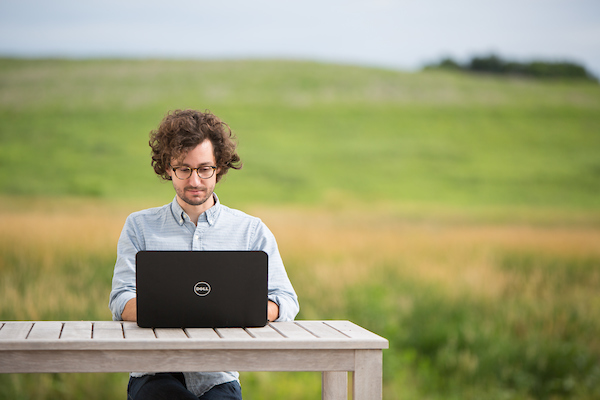 An individual sitting at a table with an open laptop with a rolling prairie behind them.